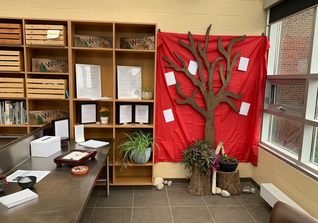 a photo of the reflection room, a table with writing supplies and a red cloth hanging on the wall with a tree and peoples' written memories and reflections on grief posted on the sheet around the tree.