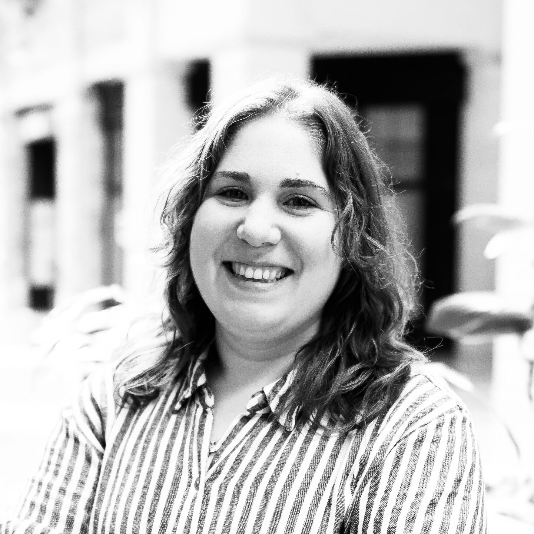 A white woman with wavy, shoulder-length brown hair in a white and grey striped shirt smiles into the camera.