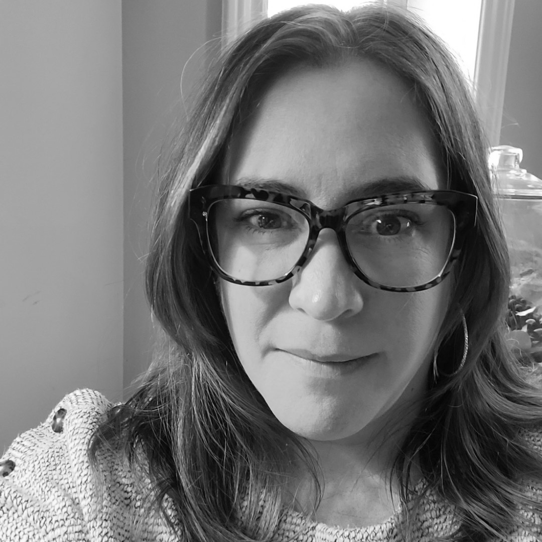 a woman with light brown hair and glasses looks into the camera with a slight smile