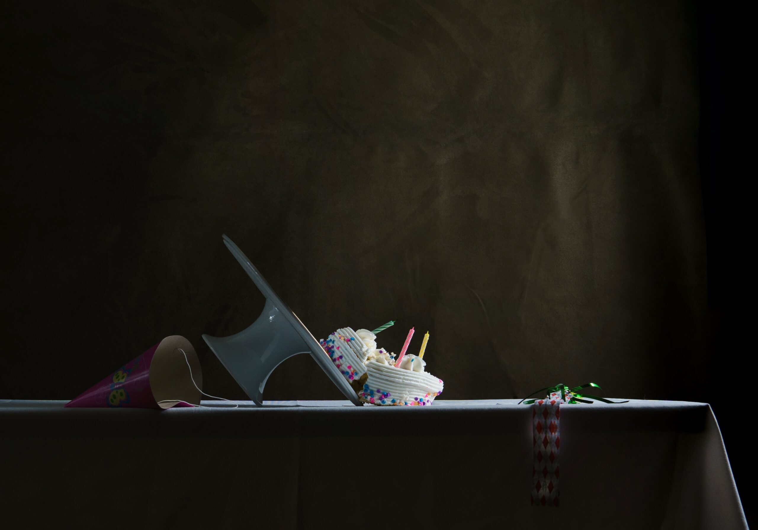 a birthday cake has fallen alone with it's cake stand. It's broken on the counter. There is a party hat in the shadows on the table behind it.